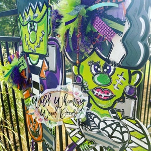 Può includere: Due appendini per porte in legno a tema Halloween con il mostro di Frankenstein. Uno è verde con un cappello viola e l'altro è verde con una camicia a righe bianche e nere. Entrambi hanno gambe a righe bianche e nere e un design a ragnatela bianca e nera. Il testo "Wild Whimsy Wood Decor" è visibile nell'immagine.
