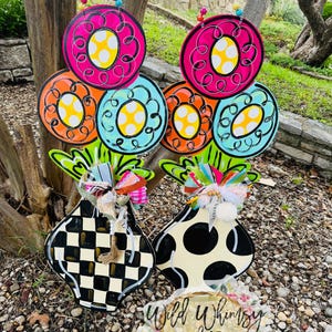 May include: Two colorful wooden flower arrangements with black and white checkered and polka dot vases. The flowers are painted in shades of pink, orange, blue, and yellow. The vases are attached to wooden stakes. The sign at the bottom reads "Wild Whimsy Wood Decor".