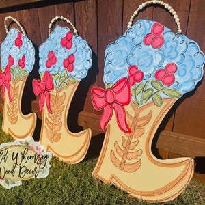 May include: Three wooden cowboy boot-shaped door hangers. Each features a light blue floral arrangement with red berries, a red bow, and a beaded handle. The boots are painted in shades of tan and brown, with the text "Wild Whimsy Wood Decor".