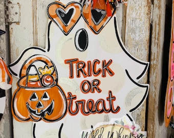 Puede incluir: Decoración de Halloween con forma de fantasma blanco con contornos negros, que lleva gafas de sol en forma de corazón. Presenta una calabaza de Halloween con caramelos y las palabras "Trick or Treat" en naranja. Un lazo con acentos de murciélago remata la decoración.