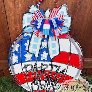 Pode incluir: Cabide de porta de madeira patriótico em forma de esfera, com um esquema de cores vermelho, branco e azul. O design inclui um laço, um picolé e o texto "Party in the USA."