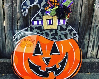 Puede incluir: Un colgador de puerta de Halloween con una calabaza con cara de jack-o'-lantern y un sombrero de bruja con estampado de leopardo. El sombrero tiene una banda de rayas moradas y blancas y una hebilla dorada. La calabaza es naranja con ojos negros, una nariz y una boca.