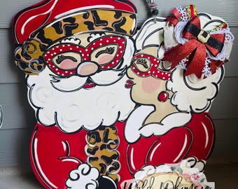 May include: A wooden door hanger featuring a painted Santa and Mrs. Claus kissing. Both wear red polka-dot glasses. Santa has a leopard print hat and trim, while Mrs. Claus has a large red and black bow. A logo reads Wild Whimsy Wood Decor.