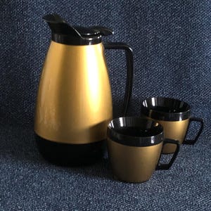 May include: A vintage gold and black coffee set. The set includes a tall, pear-shaped carafe with a black lid and handle, and two matching mugs. The carafe and mugs have a gold exterior and black accents.