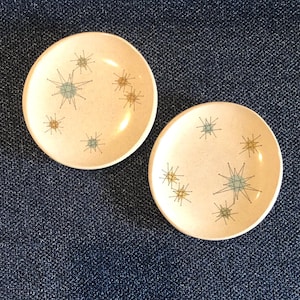Op de afbeelding: Twee kleine, ronde, crèmekleurige keramische borden met een gespikkelde afwerking. Elk bord heeft een sterrenbeeldachtig ontwerp in teal, bruin en zwart. De borden zijn waarschijnlijk bedoeld voor het serveren van kleine porties of als decoratieve items.