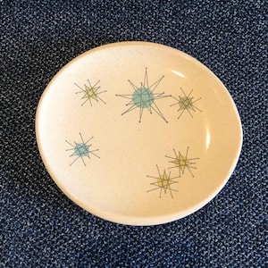 May include: A cream-colored ceramic plate with a speckled surface. The plate is decorated with starburst designs in light blue, yellow, and black. The plate has a slightly raised edge and a round shape. This plate is perfect for serving appetizers or desserts.