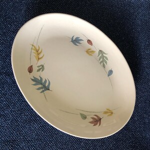 Puede incluir: Plato ovalado de color crema con diseño de hojas. Las hojas son en tonos azules, verdes, amarillos y marrones, dispuestas en un patrón circular alrededor del borde. El plato está sobre una superficie azul.