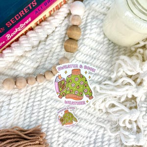 May include: Two stickers with the text "Sweater & Book Weather" and a green sweater design with flowers and a book. The stickers are on a white, textured surface with a book, wooden beads, and a candle.
