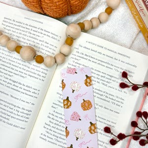 May include: A bookmark with a light purple background featuring a pattern of pumpkins in various colors and pink bows. The bookmark is placed in an open book with a pumpkin and wooden bead garland in the background.