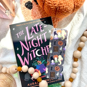 May include: A book titled "The Late Night Witches" with a matching bookmark featuring witch illustrations, pumpkins, and candles. A wooden bead garland and a crocheted pumpkin are also in the image, creating a Halloween theme.