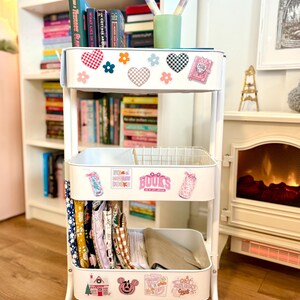 May include: A white three-tiered rolling cart with various stickers, including hearts and flowers. The top shelf holds books and a green cup with pencils. The middle shelf has a wire basket. The bottom shelf contains folded fabrics.