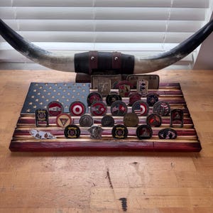 May include: A wooden American flag with red and white stripes and a blue star field, displaying a collection of challenge coins. A pair of longhorns is mounted above the flag. The coins feature various designs and emblems.