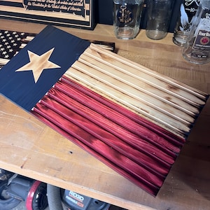 May include: Wooden Texas flag with a star and stripes. The flag is made of stained wood, with a blue canton and a gold star. The stripes are red and natural wood. The flag is displayed on a wooden surface.