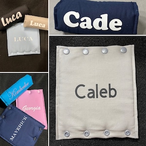 May include: Assortment of personalized baby accessories in various colors, including black, tan, light blue, navy, and pink. Each item features a name in a contrasting color. The items include a square pad with snaps and a cylindrical pillow.