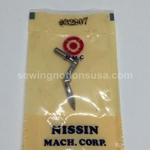 May include: A close-up of a silver sewing machine needle in a clear plastic bag with a red and white label. The label says "#32807" and "NISSIN MACH. CORP."