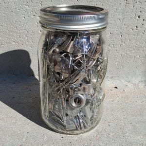 May include: A clear glass mason jar with a silver-colored metal screw-top lid, filled with a collection of assorted silver-toned metal sewing machine presser feet and various small mechanical hardware components visible through the glass.