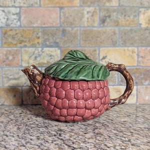 May include: A ceramic teapot shaped like a raspberry, with a textured, reddish-pink body and a green leaf-shaped lid. The handle and spout are designed to resemble tree branches. A unique and decorative kitchen item.