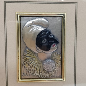 May include: A silver relief sculpture of a person wearing a white mask with a black eye mask and a gold and white ruff collar. The sculpture is framed in a gold frame and has the text "Mida 5" and "R 142 G 325" on the bottom.