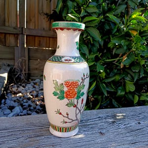 Könnte beinhalten: Eine weiße Keramikvase mit floralem Design. Die Vase hat einen grünen Rand, ein dekoratives Band und eine detaillierte Illustration von orangefarbenen Blumen und grünen Blättern. Die Vase ist etwa 25 cm hoch.