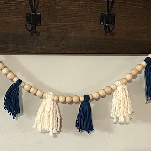 Op de afbeelding: Een witte en marineblauwe kwastengordijn met houten kralen. De gordijn is opgehangen aan een donkerbruin houten oppervlak.