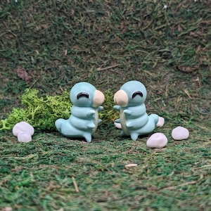 May include: Two small blue and white ceramic caterpillars facing each other. The caterpillars have smiling faces and are sitting on a bed of green moss.