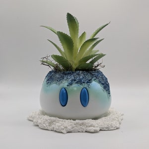 May include: A white ceramic planter shaped like a cartoon character with blue eyes. The planter is filled with a green succulent plant.