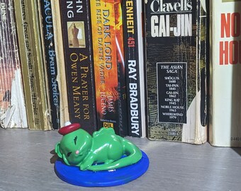 Könnte beinhalten: Eine grüne Froschfigur mit roter Mütze und blauem Sockel vor einem Bücherregal. Die Figur liegt entspannt. Auf dem Regal befinden sich Bücher mit Titeln wie "Gai-Jin" und "Fahrenheit 451".