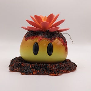 May include: A yellow and orange ceramic figurine with two black eyes and a red flower on top. The figurine is sitting on a black and orange base that resembles lava.