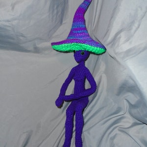May include: A handmade, purple crochet doll with a tall, pointed hat. The hat features a green brim and is striped with shades of purple, blue, and pink. The doll has a slender body, long legs, and arms. The doll is sitting.