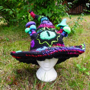 May include: A colorful, crocheted witch hat with a wide brim and a pointed top. The hat features a large, green and black eye design, with accents of purple, blue, and neon green. The hat is displayed on a white mannequin head.