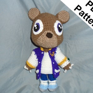 May include: A handmade crochet bear doll with large eyes, wearing a white, purple, and gold jacket, blue jeans, and blue and gray shoes. The bear is brown and tan. The text "PDF Pattern" is in the upper right corner.