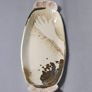 May include: A white ceramic serving dish with a hand design and brown and black splatter paint. The dish has a rounded oval shape with pink floral accents on the ends.