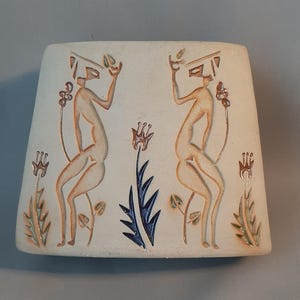 May include: A ceramic tile featuring a relief design of two figures with stylised bodies and floral motifs in shades of brown, green, blue, and white.