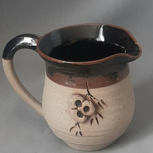 May include: A handmade ceramic pitcher with a beige body, a dark brown rim, and a black handle. The pitcher features a floral design on the front. The handle is beige with a black top. The pitcher is suitable for serving beverages.