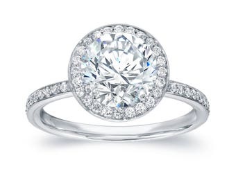 Könnte beinhalten: Ein silberfarbener Ring mit einem großen, runden, transparenten Stein im Brillantschliff, der von einem Kranz aus kleineren, runden, transparenten Steinen umgeben ist. Die Ringschiene ist schmal und mit einer Reihe kleiner, runder Steine besetzt.