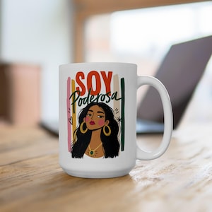 May include: A white ceramic mug featuring a graphic illustration of a person with long dark hair, gold hoop earrings, and a gold necklace. Above the illustration, the text reads SOY Poderosa in red and green letters against vertical stripes.