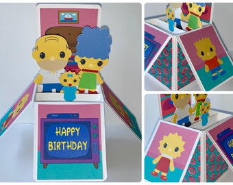 May include: A pop-up card featuring the Simpsons family. The card is open and shows Homer, Marge, Maggie, and Bart. The card has a pink background with a blue television screen that says "Happy Birthday".
