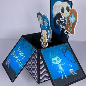 May include: A black pop-up card with a blue, white, and black chevron pattern on the inside. The card is open and has a pop-up scene with characters from the Nightmare Before Christmas and Corpse Bride movies. The card says 'Happy Birthday' on the front.