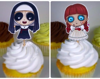 May include: Two cupcakes topped with decorative paper cutouts. One features a cartoon nun in a black habit with a red sash. The other shows a cartoon doll with red pigtails and a white dress. The cupcakes have white frosting.