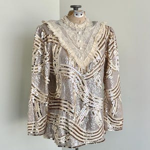 May include: A vintage silver and gold sequined top on a mannequin. The long-sleeved blouse has a high neckline with a cream lace collar and button details. The body of the top is covered in sequins arranged in a wave pattern, creating a shimmering effect.