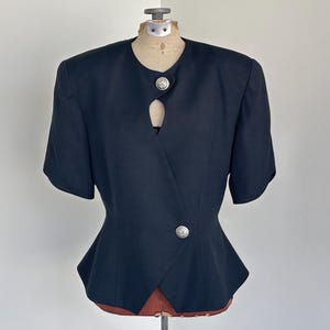 May include: A navy blue short-sleeved blazer with a unique wrap design. The blazer features two silver-colored buttons and a tailored fit. The garment is displayed on a vintage dress form, showcasing its structured silhouette.