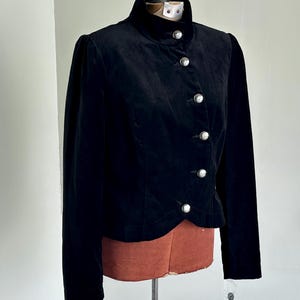 May include: A black velvet jacket featuring a high collar and silver buttons. The jacket has long sleeves and is displayed on a mannequin. The design is classic and elegant, suitable for formal wear. The jacket is made of velvet.