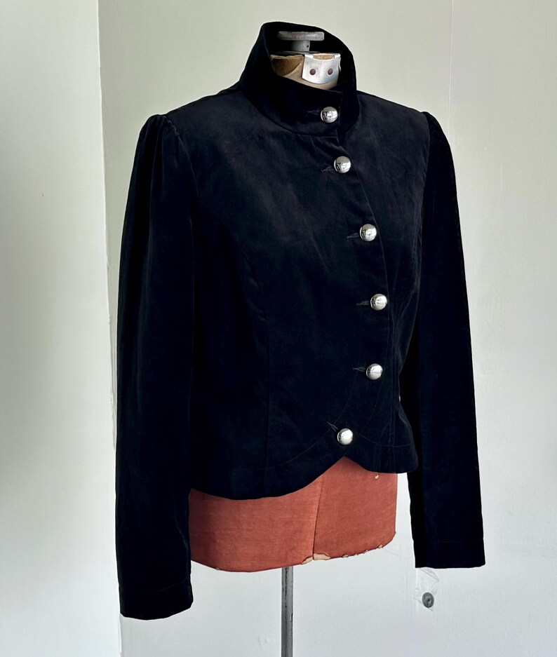May include: A black velvet jacket featuring a high collar and silver buttons. The jacket has long sleeves and is displayed on a mannequin. The design is classic and elegant, suitable for formal wear. The jacket is made of velvet.