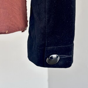May include: Detail of a black jacket sleeve with a shiny, oval button. The fabric has a soft, velvety texture. The jacket is dark, and the button reflects light.