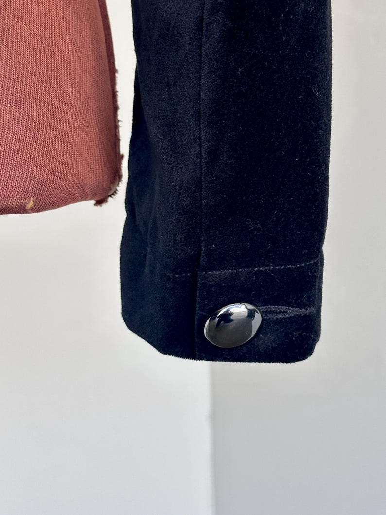 May include: Detail of a black jacket sleeve with a shiny, oval button. The fabric has a soft, velvety texture. The jacket is dark, and the button reflects light.