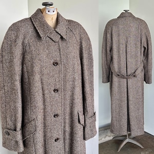 May include: A long, brown tweed coat with a classic design. The coat features a collared neckline, button-down front, and pockets. The coat is displayed on a mannequin, showcasing its full length and tailored fit. The coat has a belt in the back.