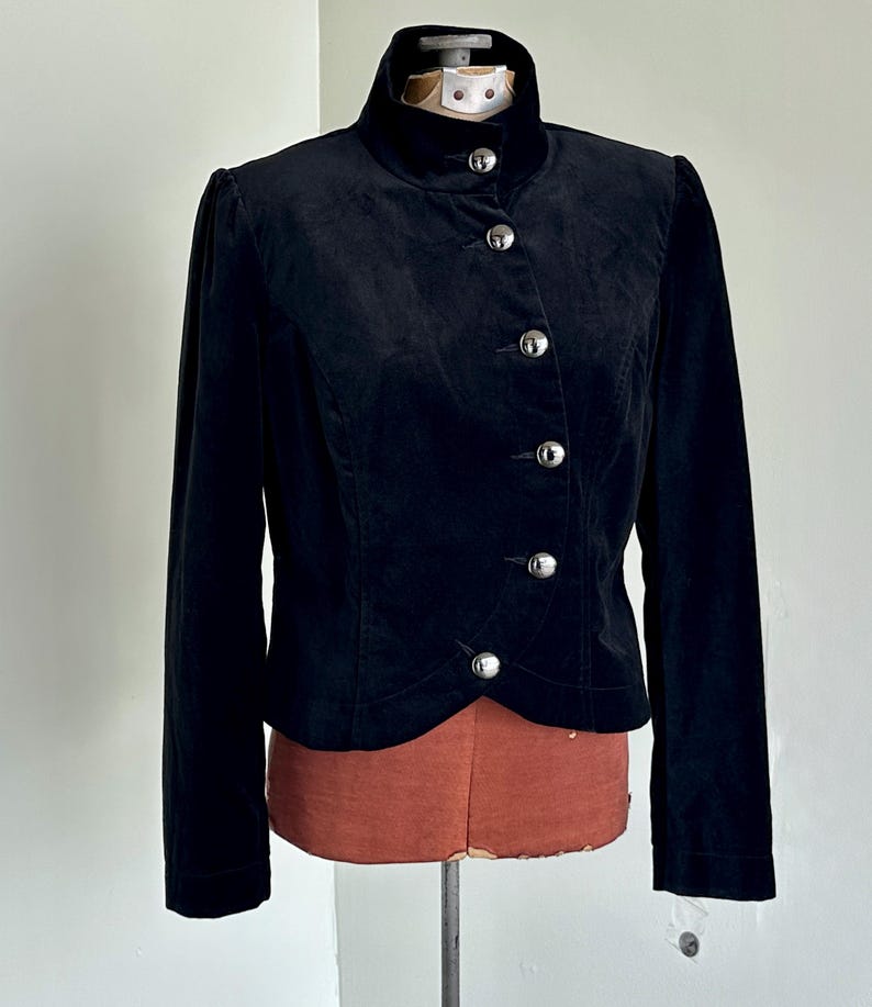 May include: A black velvet jacket with a high collar and silver buttons. The jacket has long sleeves and is displayed on a mannequin. The design is classic and elegant, suitable for formal wear. The jacket is approximately 60 cm in length.