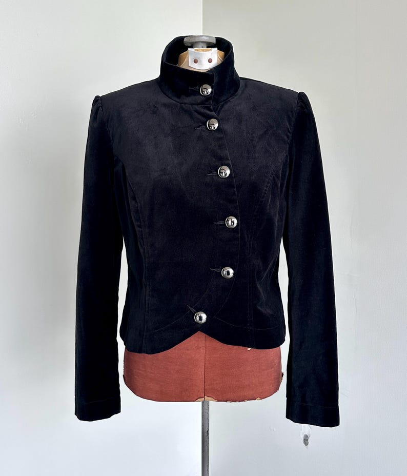 May include: A black velvet jacket with a high collar and silver buttons. The jacket has long sleeves and a tailored fit. The buttons are round and shiny. The jacket is displayed on a mannequin.