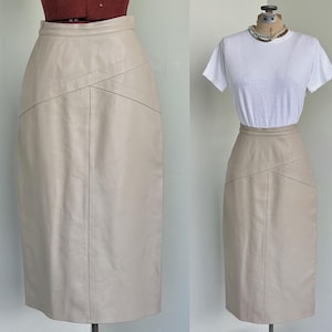 May include: A beige leather pencil skirt with a high waist and a unique angled seam design. The skirt is displayed on a mannequin, showcasing its tailored fit and classic style. A versatile piece for various outfits.