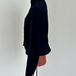 May include: A black jacket with a high collar and button details. The jacket has long sleeves and a fitted waist. The fabric appears to be a soft, velvety material. The jacket is displayed on a mannequin.